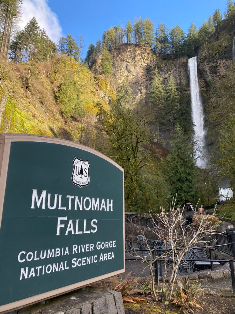 Multnomah Falls entrance sign with the waterfall and forested cliffs in the background